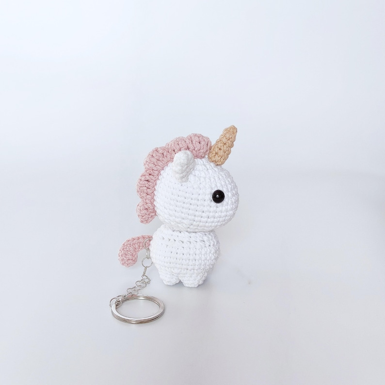 May include: White crocheted unicorn keychain with a pink mane and a gold horn. The keychain has a silver metal ring.