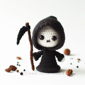 Può includere: Una bambola Grim Reaper in crochet nero con un viso bianco e grandi occhi neri. La bambola tiene un bastone marrone con una falce nera attaccata all'estremità.