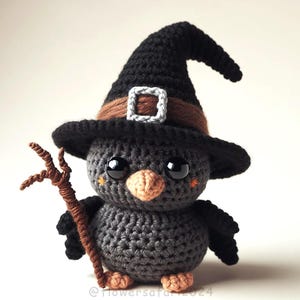 Op de afbeelding: Een gehaakte zwarte vogel die een zwarte heksenhoed met een bruine riem draagt en een bruine tak vasthoudt. De vogel heeft grote zwarte ogen en een kleine oranje snavel.