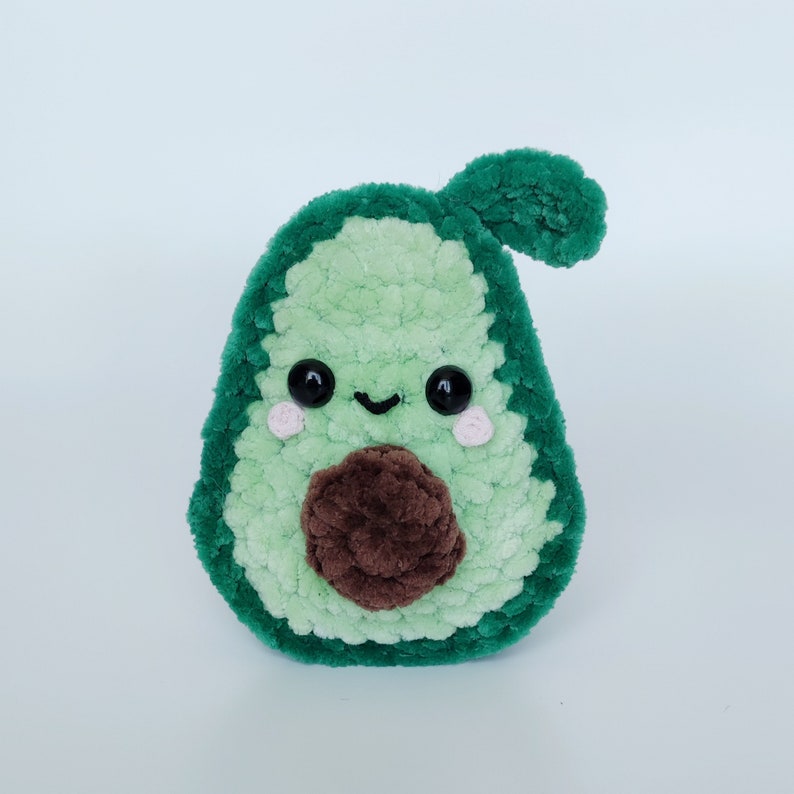 May include: A green crocheted avocado plush toy with a brown crocheted pit and a smiling face with black eyes and pink cheeks.