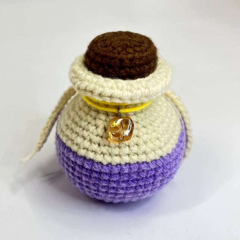 May include: A crocheted pouch in cream, purple, and brown. The pouch has a drawstring closure and a small, amber-colored charm. The top is brown, the middle is cream, and the bottom is purple. The pouch is handmade.