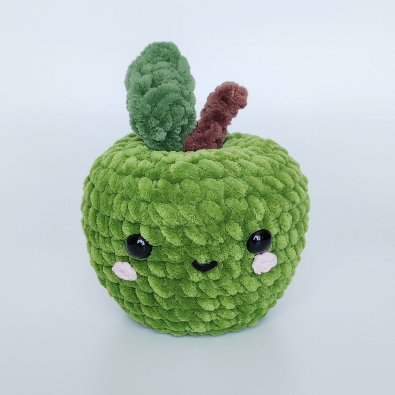 May include: A green crocheted apple plush toy with a brown stem and a cute smiling face. The apple has black eyes and pink cheeks.