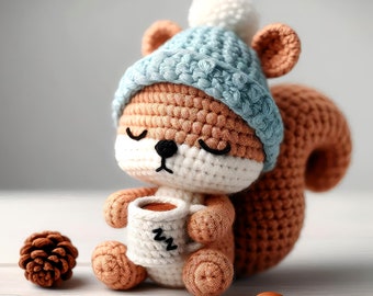 Puede incluir: Una ardilla de crochet que lleva un gorro azul y blanco con un pompón blanco, sosteniendo una taza blanca con las letras "zzz" en ella. La ardilla está sentada sobre una superficie de madera blanca.
