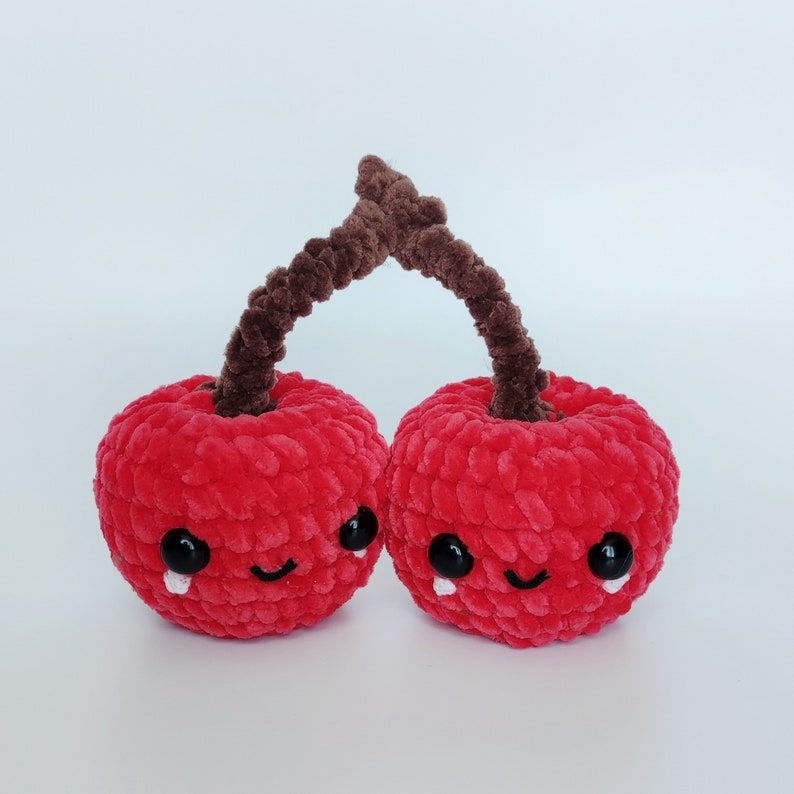 May include: Two red crocheted cherries with brown stems and smiling faces. The cherries are connected by the stems and have black eyes and white tear-shaped accents.