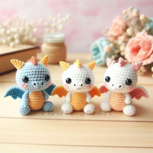 May include: Three handmade crochet dragons in blue, white, and white with orange, peach, and pink accents. Each dragon has large black eyes, small wings, and a spiky crest. The dragons are sitting on a wooden surface.