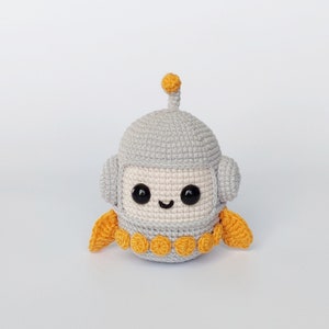 Puede incluir: Un juguete robot de crochet gris con una antena amarilla, ojos amarillos y una sonrisa amarilla. El robot tiene alas amarillas a los lados.