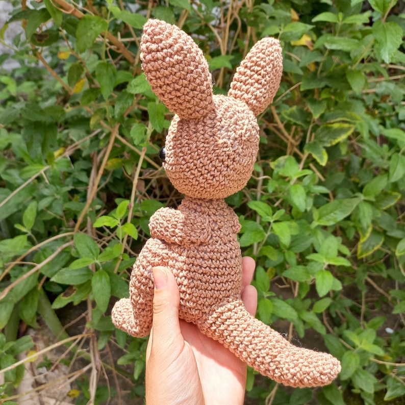 May include: A crocheted kangaroo toy in a light brown color. The kangaroo is standing on its hind legs with its tail curled behind it.