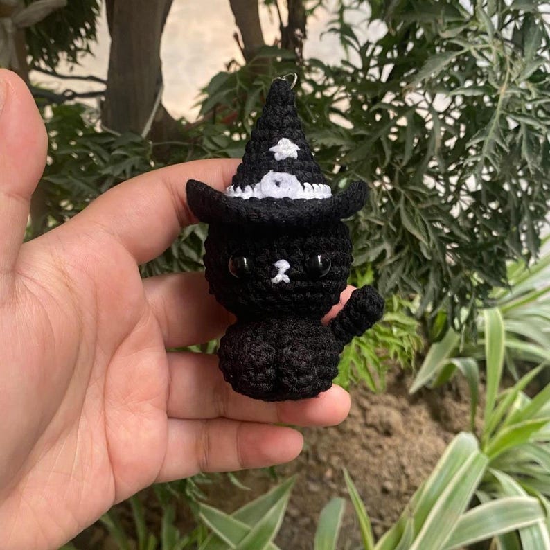 May include: A black crochet cat wearing a witch hat with white trim and a white star. The cat has a white nose and is holding up one paw. The cat is a keychain.