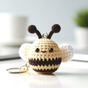 May include: A crocheted bee keychain with brown antennae, black and yellow stripes, and white wings. The bee has black eyes and a smiling mouth. The keychain is attached to a silver metal ring.
