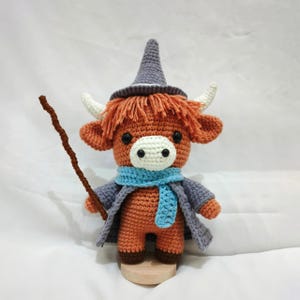 Pu&ograve; includere: Una mucca scozzese all'uncinetto marrone che indossa un cappello da strega viola, una sciarpa blu e tiene un bastone marrone.