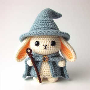 Könnte beinhalten: Gehäkeltes Häschen mit blauem Zaubererhut und Umhang, das einen braunen Stab hält. Das Häschen hat schwarze Augen und cremefarbenes Fell mit orangefarbenen Ohren. Ein goldfarbener Verschluss sichert den Umhang. Ein handgefertigtes, skurriles Design.