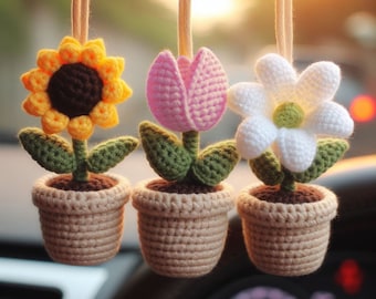 May include: Three crocheted flower pot ornaments hanging from strings. The flowers are a yellow sunflower, a pink tulip, and a white daisy. The pots are a light brown color.