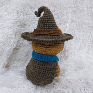 Op de afbeelding: Een gehaakt knuffeldier met een bruine hoed en een blauwe sjaal. Het speelgoed zit op een wit, pluche oppervlak.