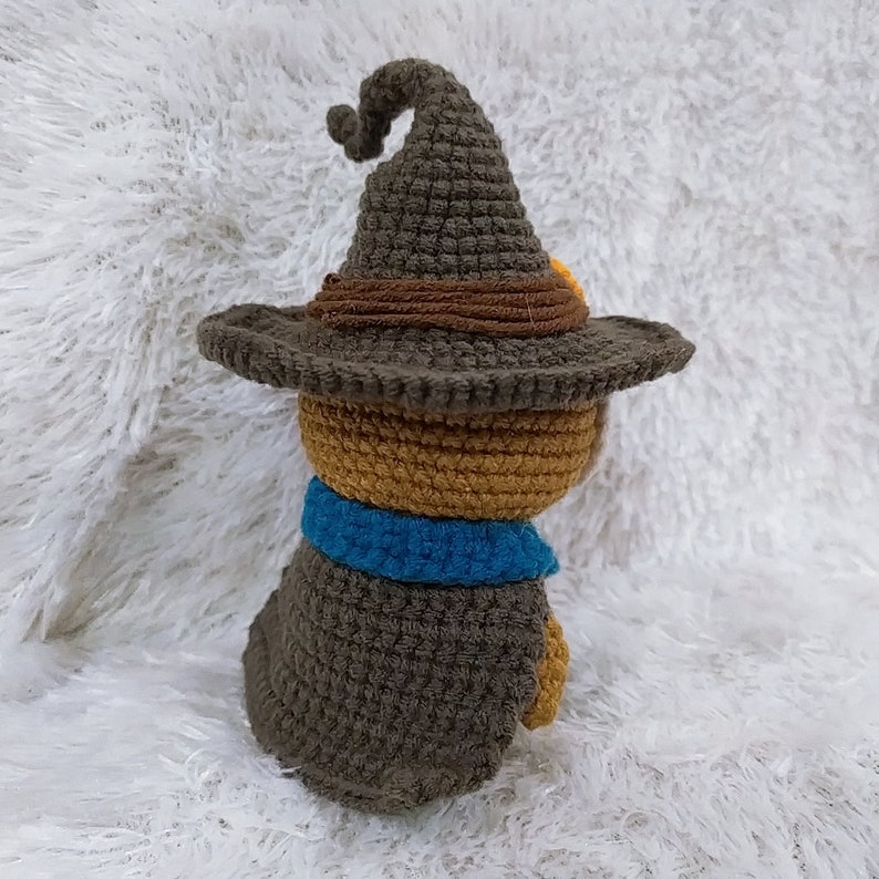 Op de afbeelding: Een gehaakt knuffeldier met een bruine hoed en een blauwe sjaal. Het speelgoed zit op een wit, pluche oppervlak.