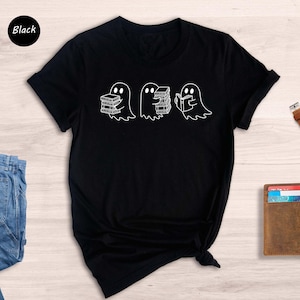 May include: Black t-shirt featuring a graphic of three ghosts interacting with books. One ghost carries a stack of books, another holds a single book, and the third ghost appears to be reading a book. The design is simple and minimalist, with white outlines on a black background. The word 'Black' is visible in the upper left corner, indicating the shirt's color.