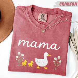 a pink t-shirt with a design of a duck and two baby ducks, along with the word "mama" written on it. The t-shirt is placed on a white surface, and there is a brown hat and some dried leaves nearby.