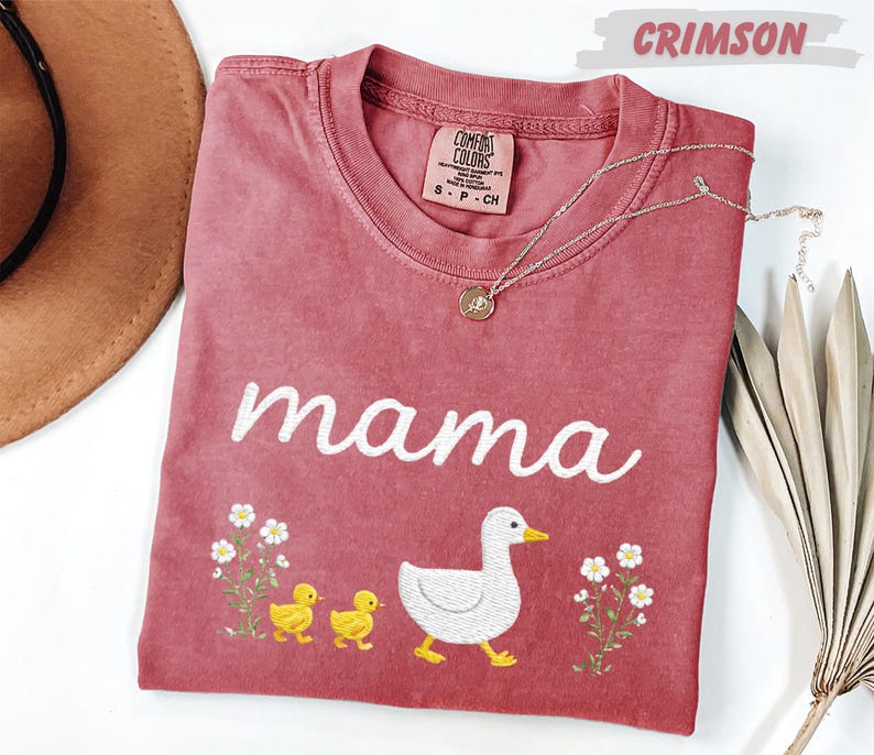 a pink t-shirt with a design of a duck and two baby ducks, along with the word "mama" written on it. The t-shirt is placed on a white surface, and there is a brown hat and some dried leaves nearby.