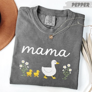 A gray t-shirt with the word "mama" printed on it, along with a small duck and flowers, is displayed on a white surface.