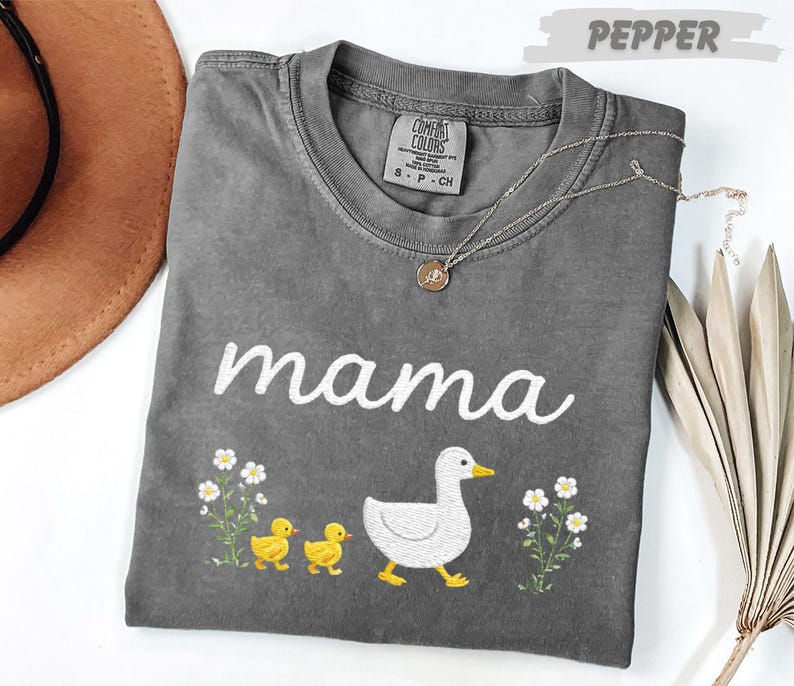 A gray t-shirt with the word "mama" printed on it, along with a small duck and flowers, is displayed on a white surface.