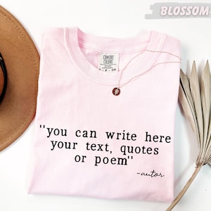 A pink t-shirt with a quote printed on it, along with a brown hat and a palm frond, all placed on a white background.