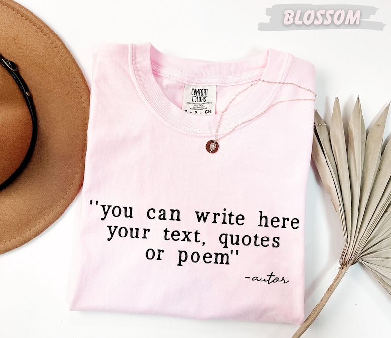 A pink t-shirt with a quote printed on it, along with a brown hat and a palm frond, all placed on a white background.