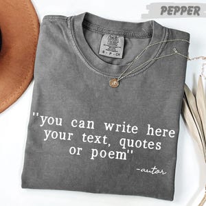 A gray t-shirt with the text "you can write here your text, quotes or poem" printed on it, along with a small pendant necklace. The t-shirt is placed on a white surface, and there is a brown hat and some dried leaves nearby.