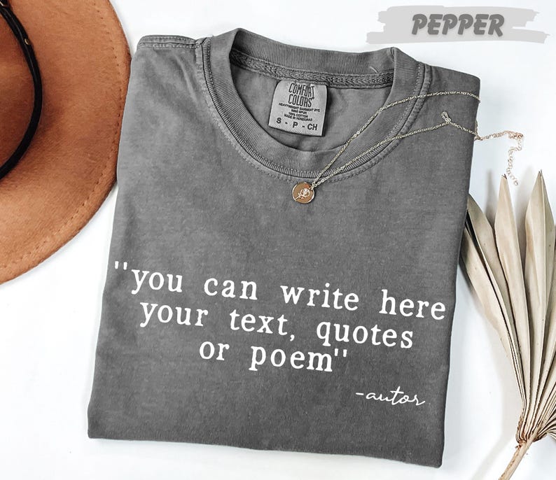 A gray t-shirt with the text "you can write here your text, quotes or poem" printed on it, along with a small pendant necklace. The t-shirt is placed on a white surface, and there is a brown hat and some dried leaves nearby.