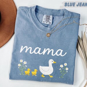 A blue t-shirt with the word "mama" and a duck and baby duck embroidered on it, along with flowers, is displayed on a white surface.