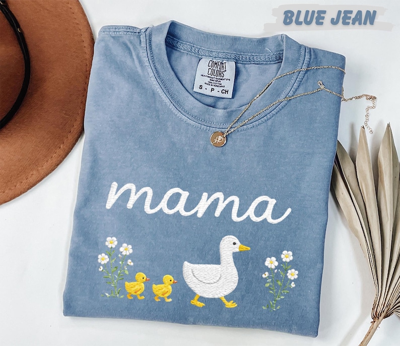 A blue t-shirt with the word "mama" and a duck and baby duck embroidered on it, along with flowers, is displayed on a white surface.