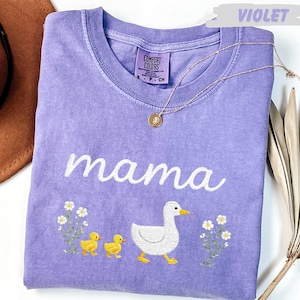 a purple t-shirt with a white text design and a small white flower design, along with a small white flower design and a small white flower design. The t-shirt is placed on a white surface, and there is a brown hat and a plant in the background.