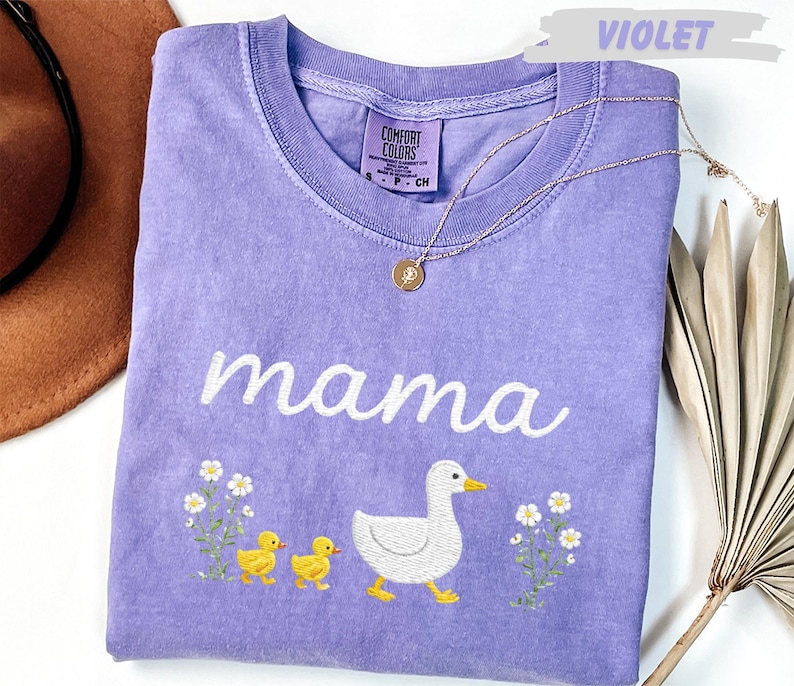 a purple t-shirt with a white text design and a small white flower design, along with a small white flower design and a small white flower design. The t-shirt is placed on a white surface, and there is a brown hat and a plant in the background.
