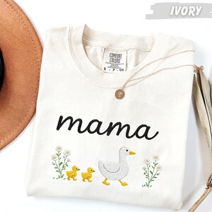 A white t-shirt with the word "mama" printed on it, along with a small duck and two ducklings, is displayed on a white background.