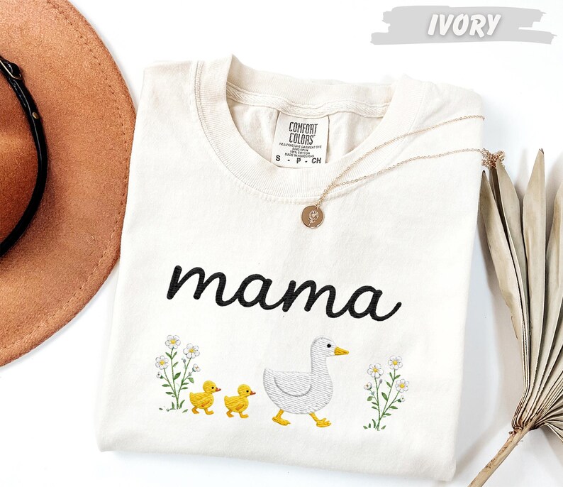 A white t-shirt with the word "mama" printed on it, along with a small duck and two ducklings, is displayed on a white background.