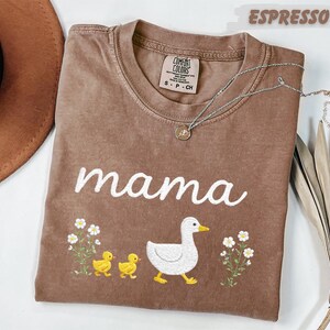 a brown t-shirt with a white duck and two yellow ducklings, along with the word "mama" written on it. The t-shirt is placed on a white surface, and there is a brown hat and a plant nearby.