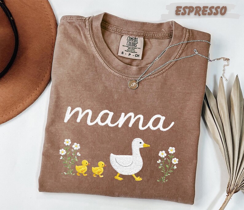a brown t-shirt with a white duck and two yellow ducklings, along with the word "mama" written on it. The t-shirt is placed on a white surface, and there is a brown hat and a plant nearby.