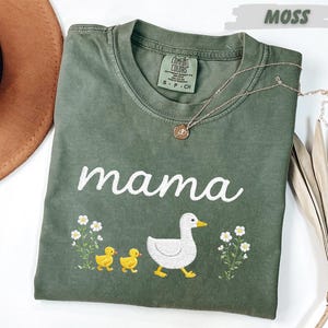a green t-shirt with a white text design and a small embroidered image of a duck and its ducklings. The t-shirt is placed on a white surface, with a brown hat and some dried leaves nearby.