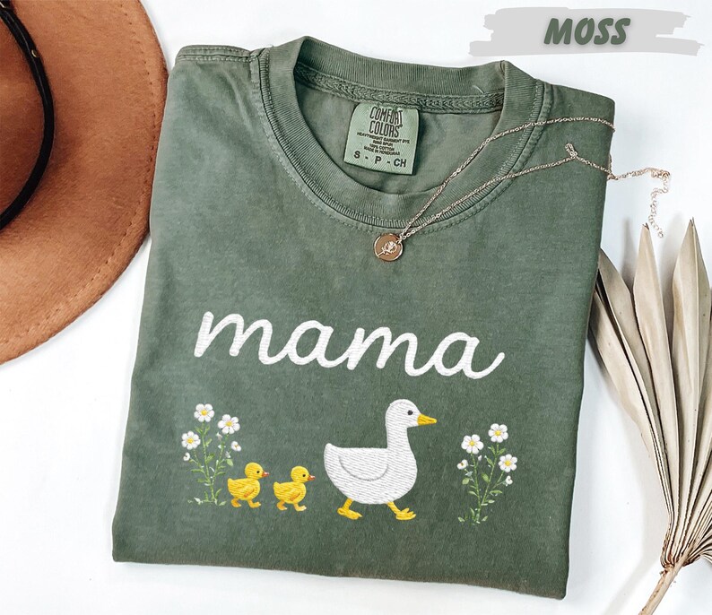 a green t-shirt with a white text design and a small embroidered image of a duck and its ducklings. The t-shirt is placed on a white surface, with a brown hat and some dried leaves nearby.