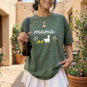 A woman wearing a green t-shirt with a graphic of ducks and a mother duck, standing in front of a building with potted plants.