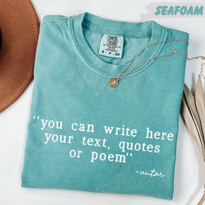 A turquoise t-shirt with a quote printed on it, along with a brown hat and some dried leaves, all placed on a white surface.