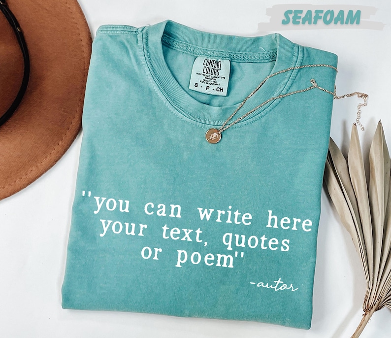 A turquoise t-shirt with a quote printed on it, along with a brown hat and some dried leaves, all placed on a white surface.