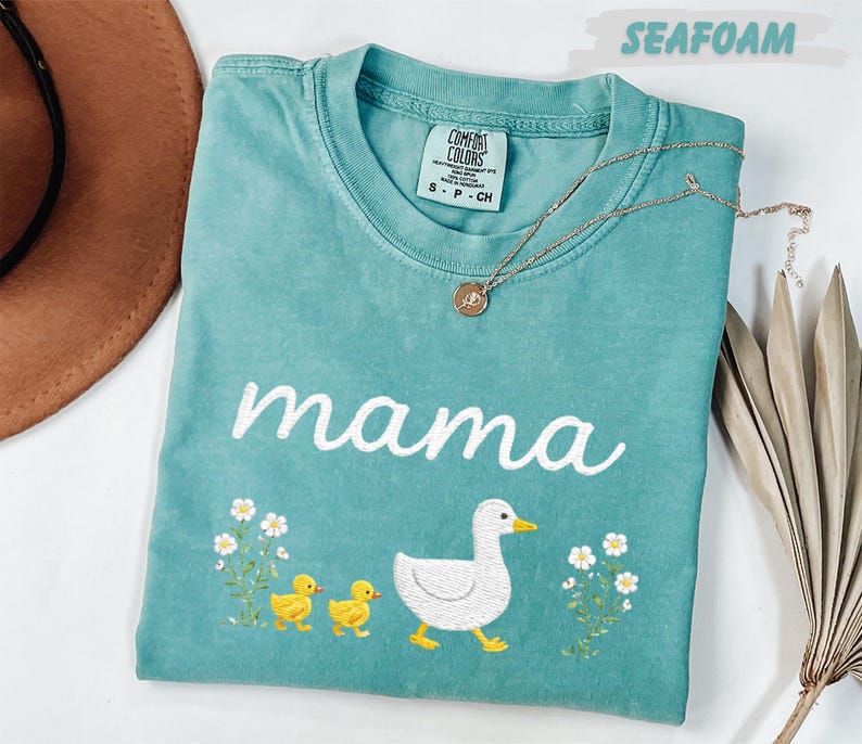 A turquoise t-shirt with a mother duck and her ducklings printed on it, along with a necklace and a hat.