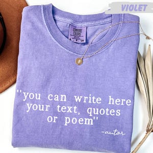 A purple t-shirt with the text "you can write here your text, quotes or poem" printed on it, along with a small gold medallion. The t-shirt is placed on a white surface, and there is a brown hat and a plant in the background.