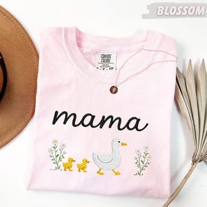 A pink t-shirt with the word "mama" and a graphic of a duck and two ducklings, along with some flowers, is displayed on a white surface.