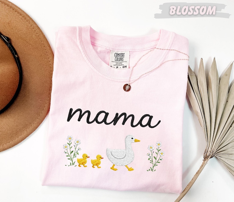 A pink t-shirt with the word "mama" and a graphic of a duck and two ducklings, along with some flowers, is displayed on a white surface.