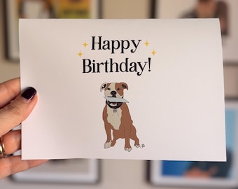 May include: A white rectangular greeting card featuring the black text Happy Birthday! centered at the top with four small yellow star icons. Below the text is a brown and white illustration of a dog wearing a black collar and holding a gray object.