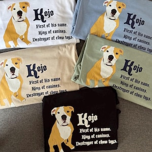 Pode incluir: Cinco camisetas diferentes com uma ilustração de um cão castanho e branco e o texto "Koja" numa fonte preta estilizada. O texto abaixo do cão diz "First of his name. King of canines. Destroyer of chew toys."