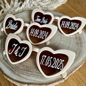 May include: Five pairs of heart-shaped sunglasses with white text on a light pink frame. The text on the sunglasses reads "Bride", "Team Bride", "14.09.2024", "J&J", and "17.05.2025".