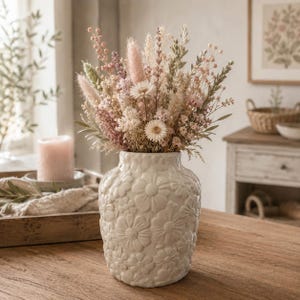 May include: A white ceramic vase with a raised floral pattern filled with a bouquet of dried flowers in shades of pink, cream, and green. The arrangement includes fluffy plumes, small daisy-like blossoms, and various textured grasses and stems.