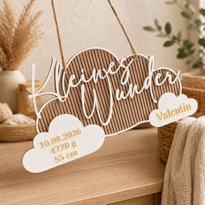 May include: A hanging wooden sign shaped like clouds with vertical slats, featuring the white text "Kleines Wunder". Attached white cloud cutouts display the name "Valentin" and birth details: "10.08.2026", "4720 g", and "55 cm" in gold.