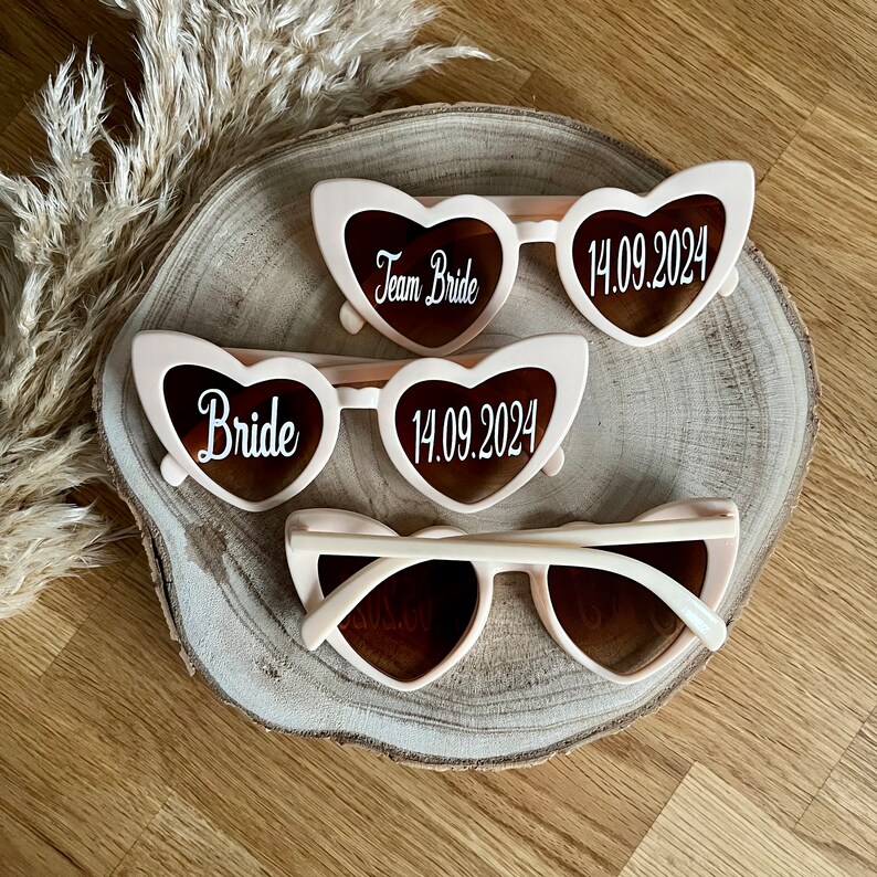 May include: Three pairs of pink heart-shaped sunglasses with white text. The sunglasses say "Team Bride", "Bride", and "14.09.2024".